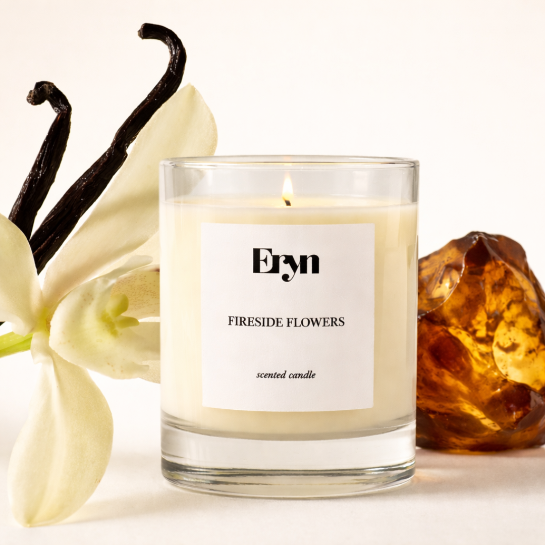 A lit FIRESIDE FLOWERS clean burning candle by Eryn surrounded by vanilla, a vanilla flower and amber on a neutral background 